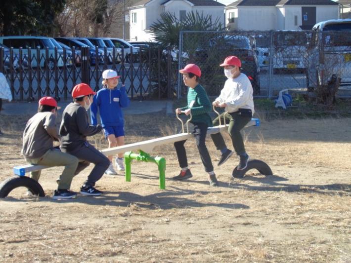 校庭にあるシーソーで遊ぶ子どもたち。2人ずつ左・右にのっており、真ん中で見ている子どもが1人いる。