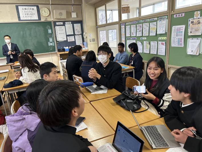 スマートフォンを見せて説明をする外国出身者とそれを聞く中学生