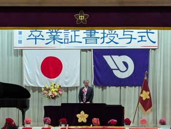 卒業証書授与式と書かれた看板の下からあいさつをする男性