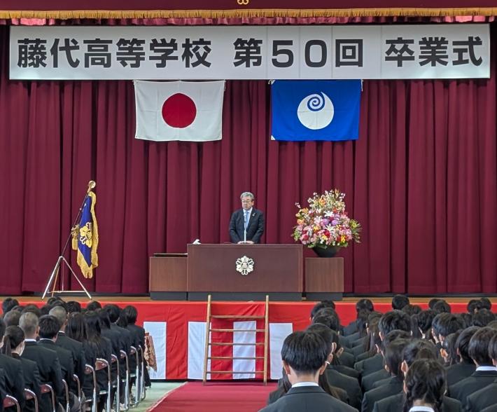 第50回卒業式と書かれた看板の下からあいさつをする男性