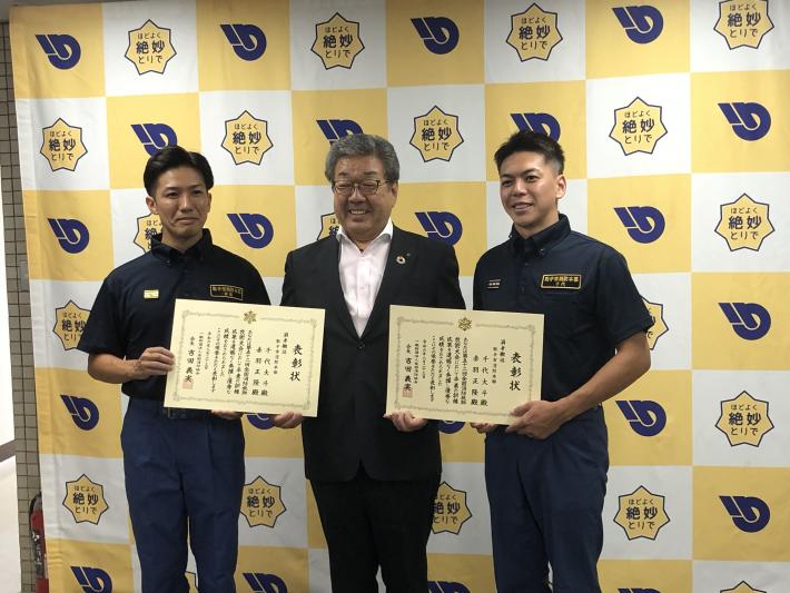 制服を着た消防職員2名とスーツを着た男性がパネルの前に立っている