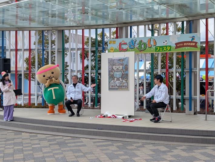 ステージ上にある絵画を隔てて2人の男性がいすにすわっている