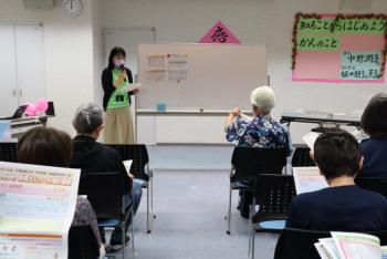 ホワイトボード横に立っている女性が座っている5名に話している