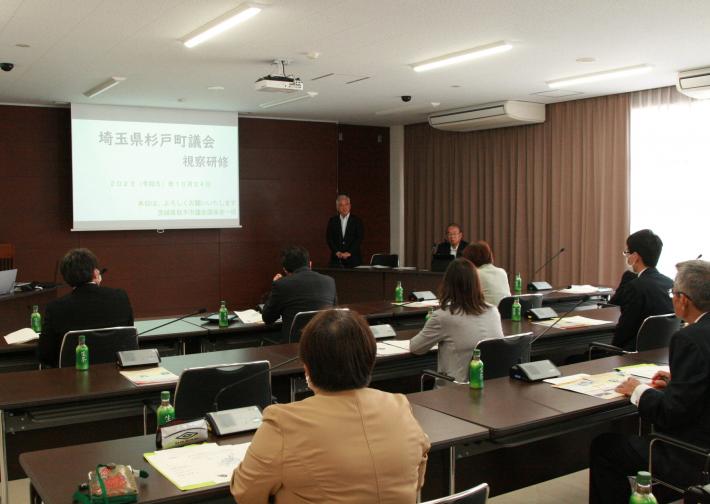 会議室の長机に男女が座り正面のスクリーンに映し出された資料を見ている