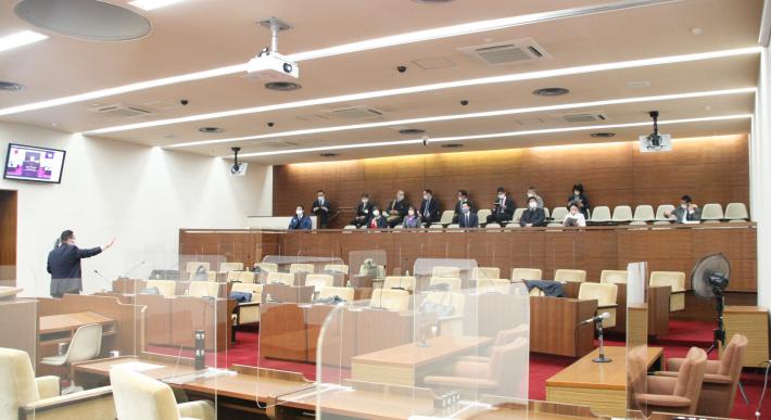 議場の傍聴席に15人の男女が座り、モニターの下にいる議会事務局職員からオンライン議案説明について説明を受けている。