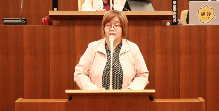 議場の壇上に、ピンク色のスーツを着た短髪の女性がこちらを向いて立っている