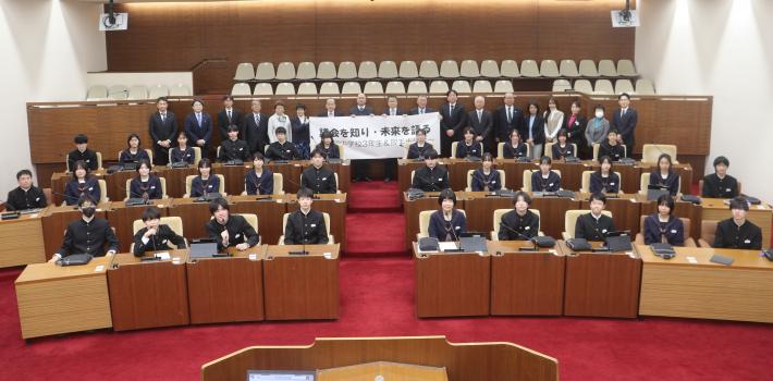 議場にたくさんの生徒が座り、その奥に議員が一列に並んで、笑顔でこちらを向いている