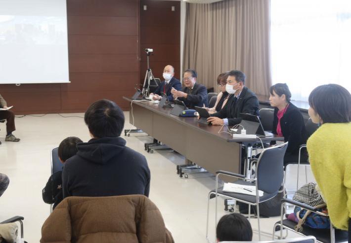 会議室で机に男性と女性の議員が座っており、その周りの椅子に参加者が座っている