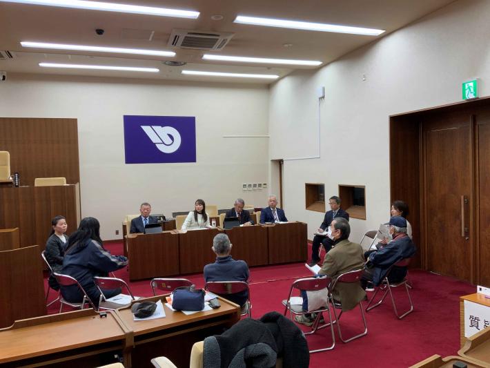 議会議場の座席に男性と女性の議員が座っており、その周りに参加者が椅子に座っている