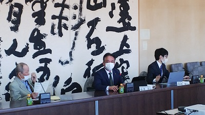 横一列の机に3人の男性が座っている。中央の男性議員(金澤委員長)が着座のまま挨拶をしている写真