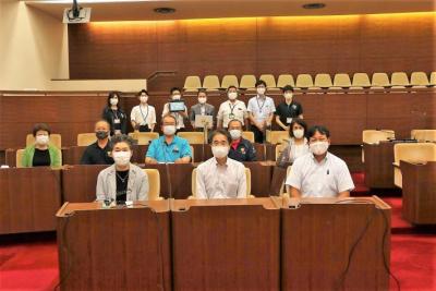 議場で発表会に参加した議員と議会事務局職員の集合写真。着席している8人の議員と後ろに一列に並んで立っている7人の事務局職員。