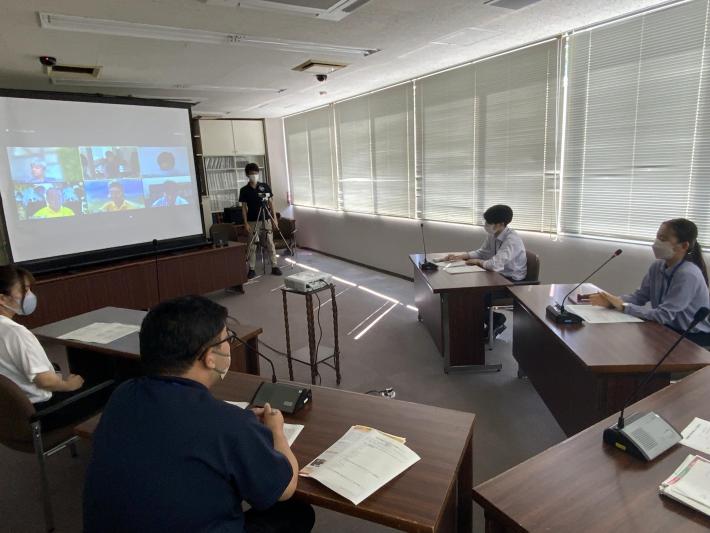 執行部控室内にいる新規採用職員がスクリーンに映る議員と対話している