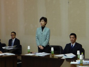 女性委員長(阿部委員長)が起立して挨拶をしている様子。両隣に男性議員および市職員が着席している。