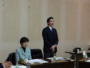 男性副委員長(関川副委員長)が起立して挨拶している。左隣に女性委員長(阿部委員長)が着席している。