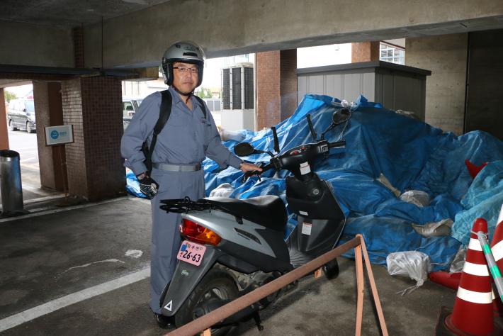 コンクリートの上にバイクを停めて、作業着を着てヘルメットをかぶった男性が立っている。