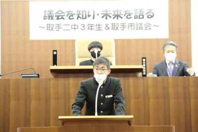 演壇で所信表明する男性生徒(小林君)。後ろには男性臨時生徒議長と男性議会事務局長が座る。