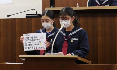 演壇で説明する共生党の女子生徒2人。右の生徒がマイクに向かって話し、左の生徒がメリットと書かれたフリップを持つ。