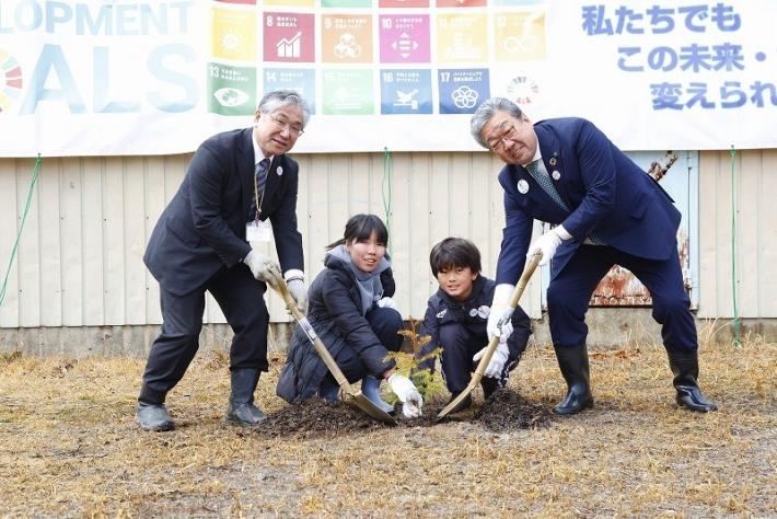 屋外にて植樹の様子。しゃがんで木を持つ児童2人と両脇に市長と校長がスコップで掘る様子。背景にSDGsの横断幕