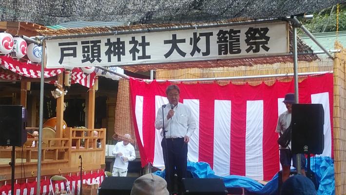 戸頭神社大灯籠まつりでの市長あいさつの様子。やぐら上に戸頭神社大灯籠祭の看板と紅白幕をバックに挨拶する男性