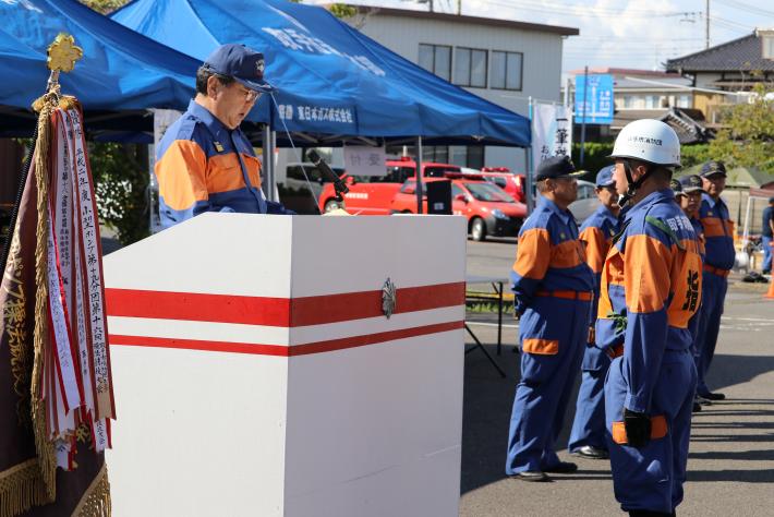 駐車場にて演台上で話をする制服をきた市長。演台下に制服を着た団員