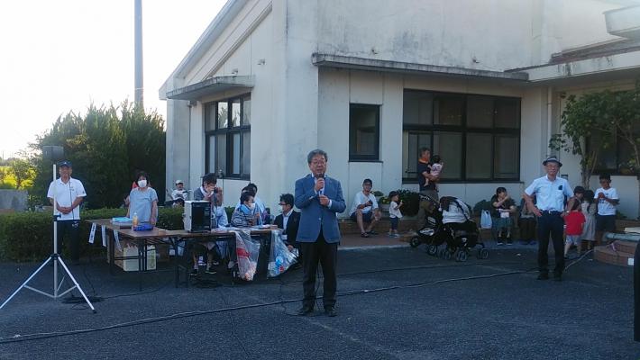 相馬地区夏まつり盆踊り大会。白壁の建物前の駐車場様の場所で男性が挨拶をしている。背後には参加者らしい人が着座。