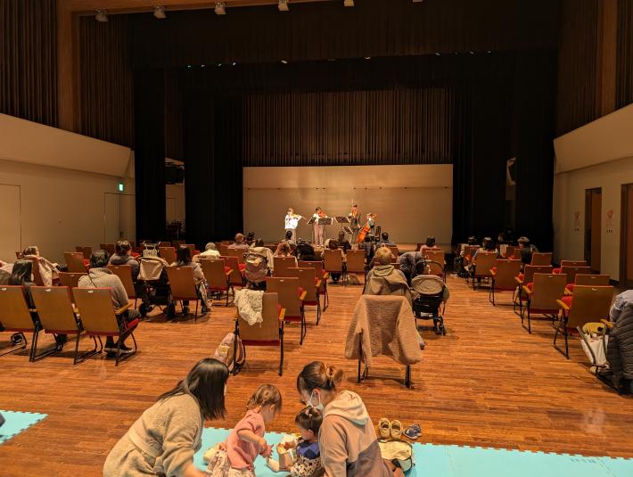 スタッキングチェアに座って演奏を聴く観客と後ろで子どもの世話をする大人