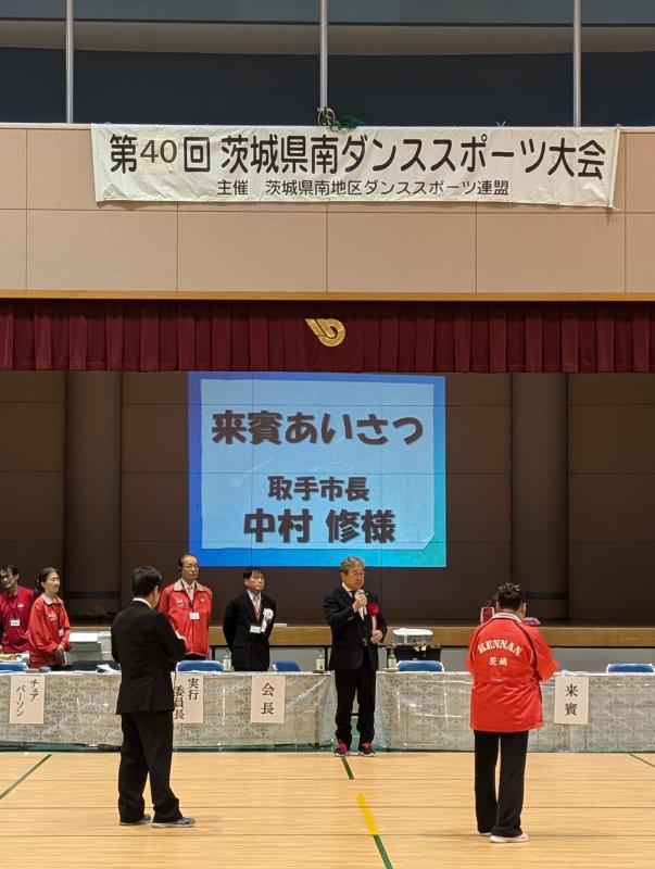 茨城県南ダンススポーツ大会と書かれた看板の下からあいさつをする男性