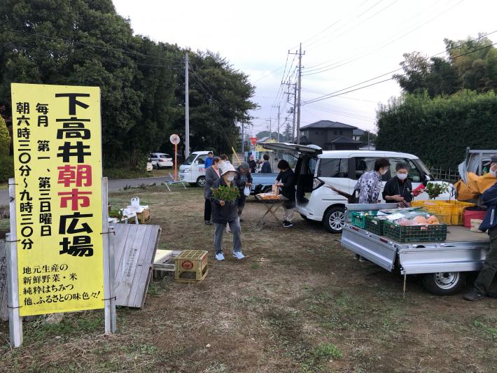 トラックの荷台にある野菜等を見る人たち。手前には下高井朝市と書いてある立て看板がある。