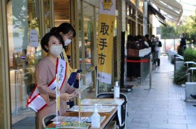 店頭のひさしの下のテーブルでたすきをかけて冊子を配る若い女性の画像