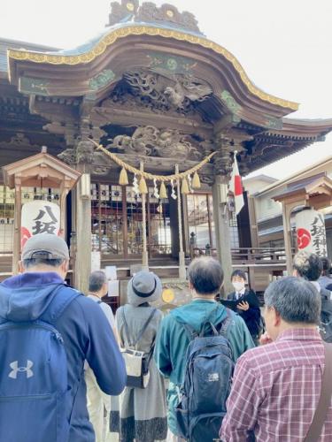 神社の境内の前で何かを説明する若い男性とそれを手前で聞いている人々の様子。