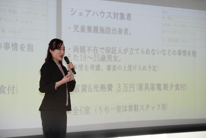 辻川百華さん発表の様子。黒スーツ姿の女性がスクリーンの前で、マイクを片手に発表を行っている。