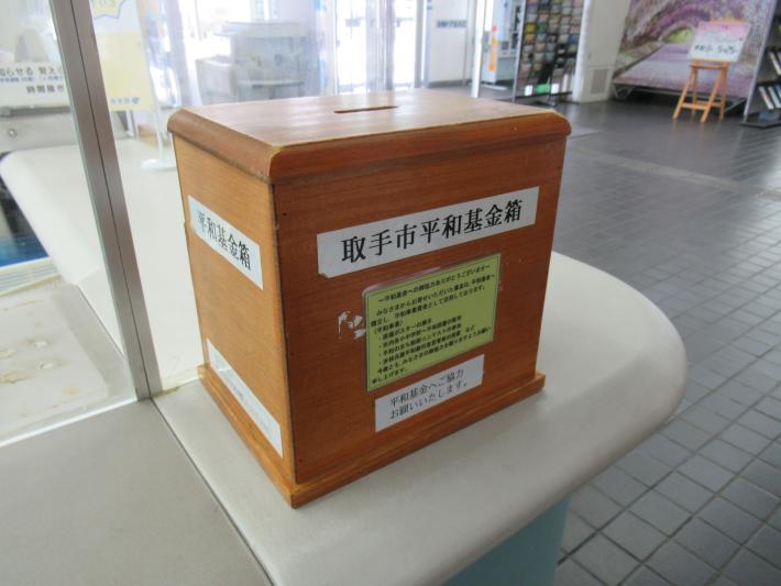 白色の受付カウンター上に置かれた木製の茶色い平和基金箱