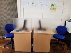 デスクが2台向かってならび、青い事務椅子が設置されている。机上には目隠しのパーティションと端末がある。